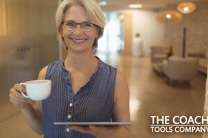 Smiling coach wearing glasses holding cup of coffee and tablet in office, relaxing while working